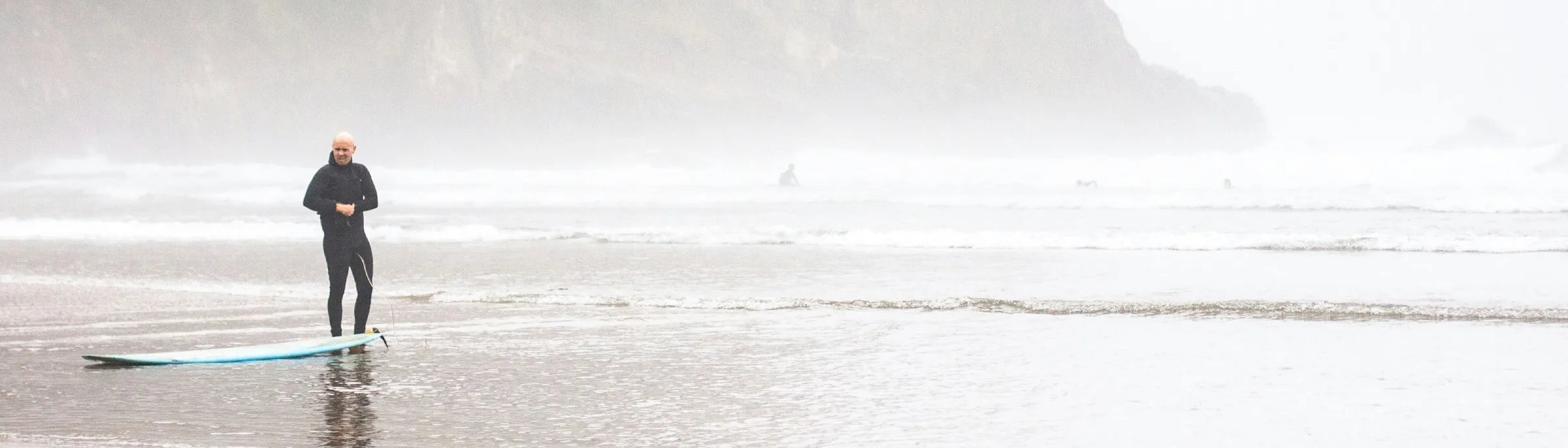 A surfer in a black steamer wetsuit stands on a misty, cold-water beach with their surfboard, ready to enter the ocean.