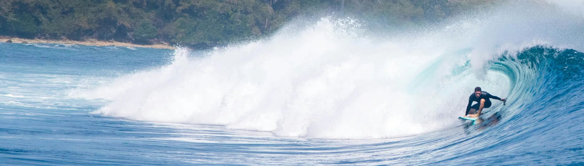 Surfer skillfully riding inside a large, barreling ocean wave with a tropical coastline in the background.