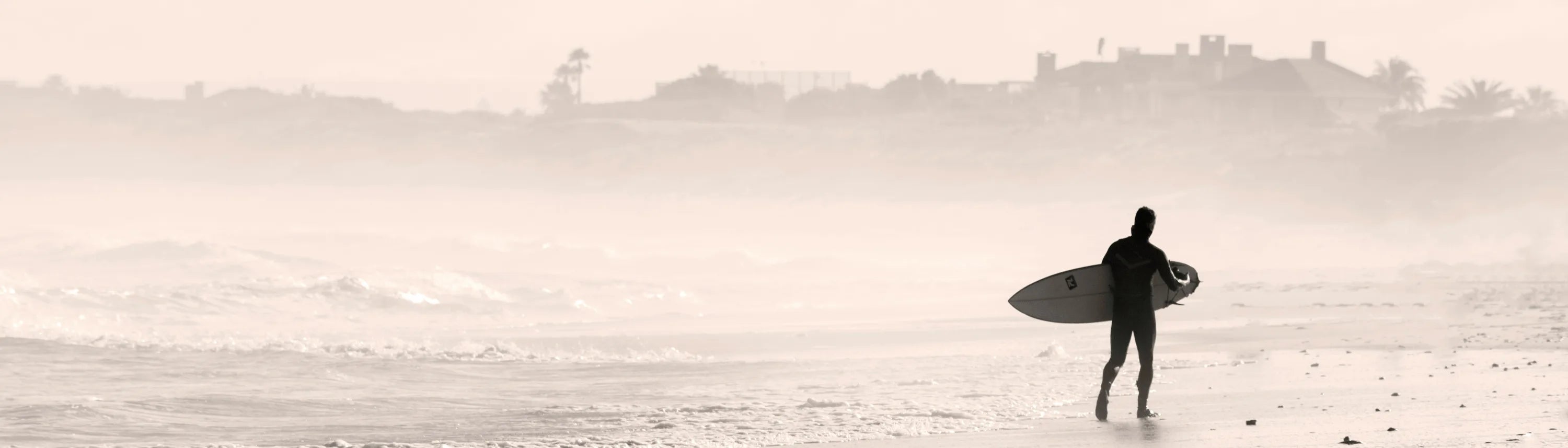 A surfer walks along a sandy beach carrying a surfboard, with the ocean in the background.